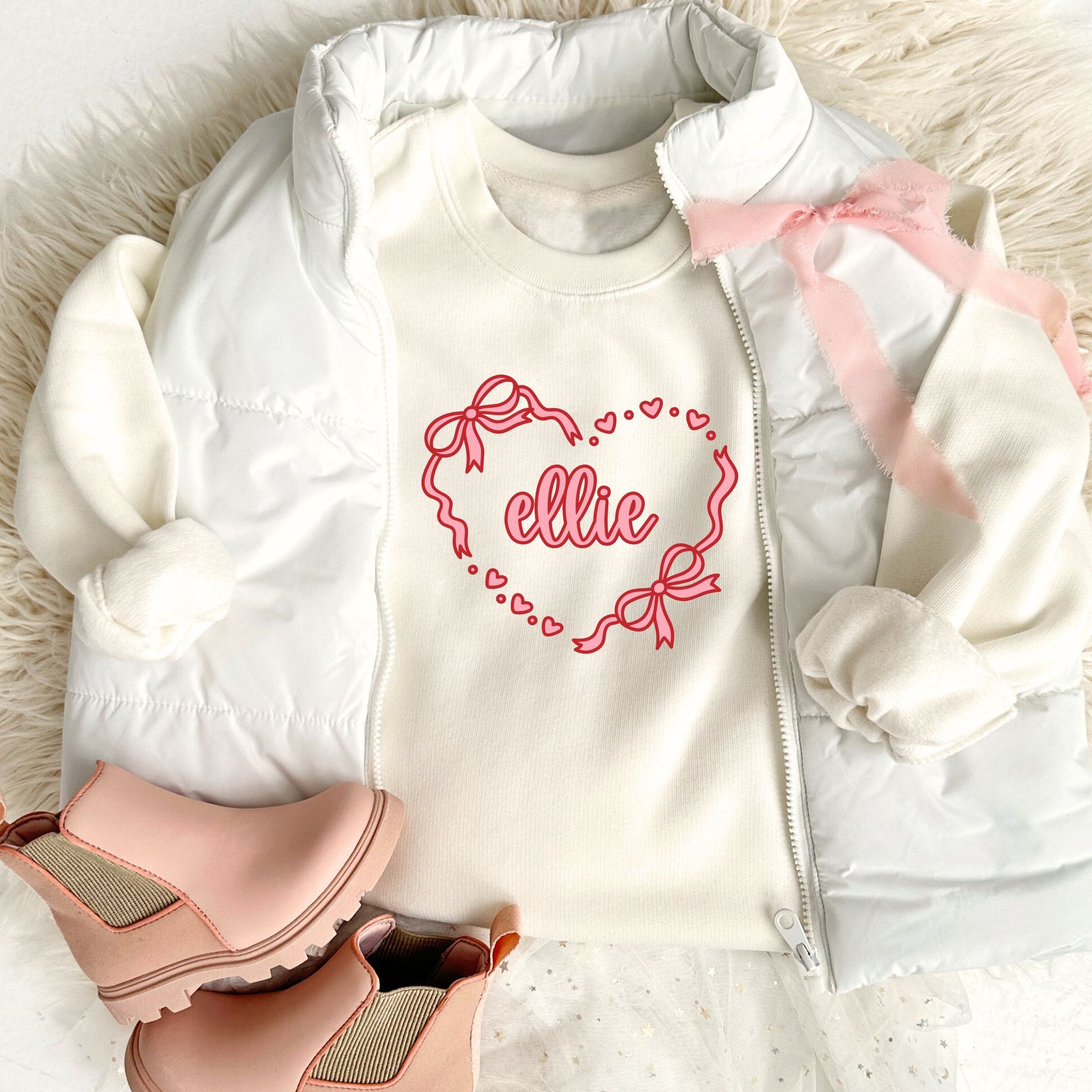 natural crewneck sweatshirt with a bow heart name printed design on the center chest, styled with a white puffer vest, pink boots, and a pale pink bow ribbon