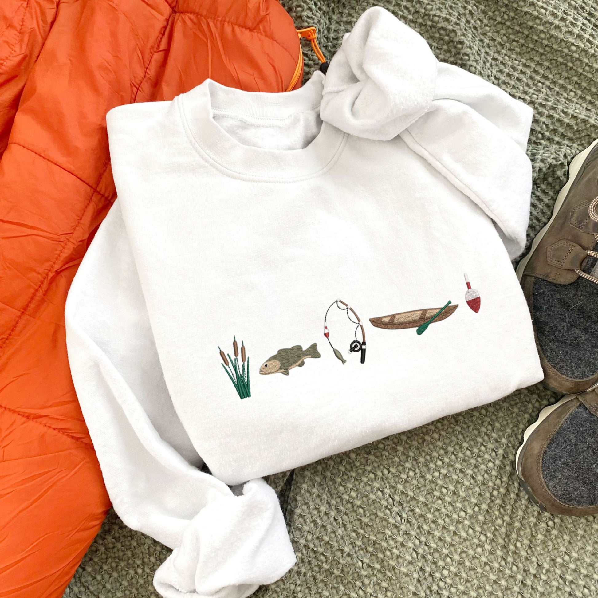 white crewneck with custom embroidered fishing icons including cattails, bass fish, fishing rod, canoe, and bobbin across the chest