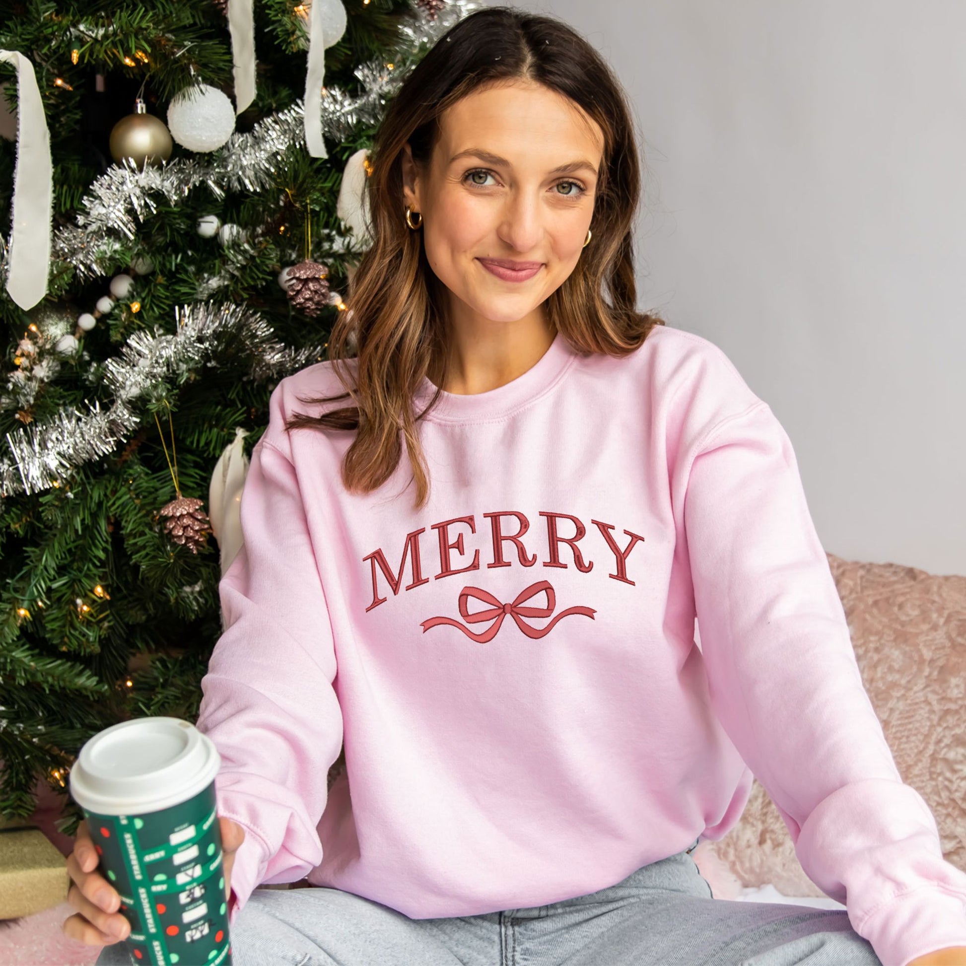 woman wearing a light pink crewneck sweatshirt with large embroidered merry and bow design in red thread across the chest