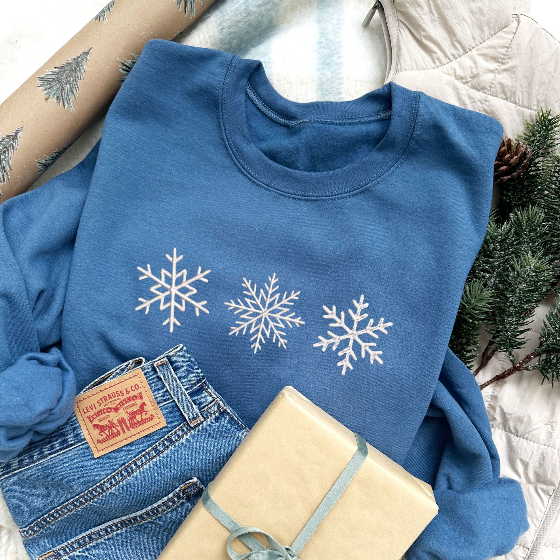 Christmas styled flat lay photo an indigo crewneck sweatshirt with3 embroidered snowflakes across the chest