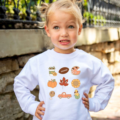 Little girl wearing a white crewneck sweatshirt with cute fall icons like a football, pumpkin, leaves, turkey leg, truck, and sunflower