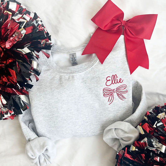 flat lay of an ash crewneck sweatshirt with embroidered name and striped bow on the left chest in white, grey, and red threads