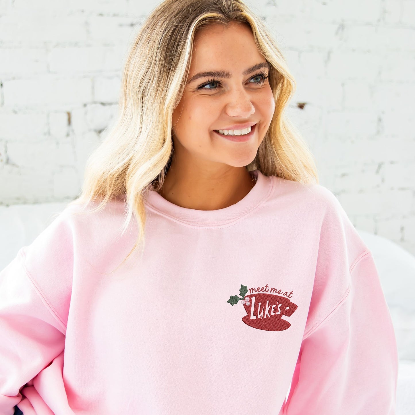 young woman wearing a light pink crewneck sweatshirt with custom embroidered "meet me at Luke's" coffee cup holly design on the left chest