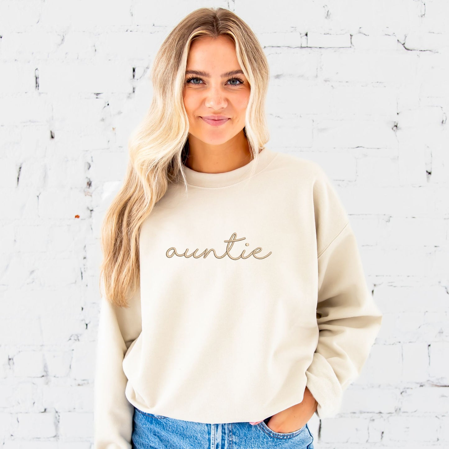 young woman wearing a sand crewneck sweatshirt with embroidered auntie in lowercase script font across the chest in camel thread