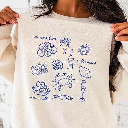 close up of girl wearing a sweet cream crewneck sweatshirt with  a printed design featuring an italian "mangia bene, ridi spesso, ama molto" and hand-drawn looking icons such as tomatoes, sardines, crab, olives, wine, and flowers