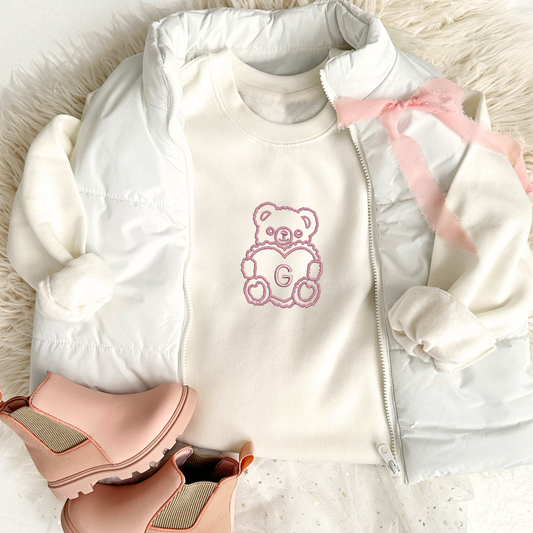 natural crewneck sweatshirt with embroidered teddy bear heart initial design on the center chest in mauve pink thread. sweatshirt is styled with a white puffer vest, pink bow, and pink ankle boots