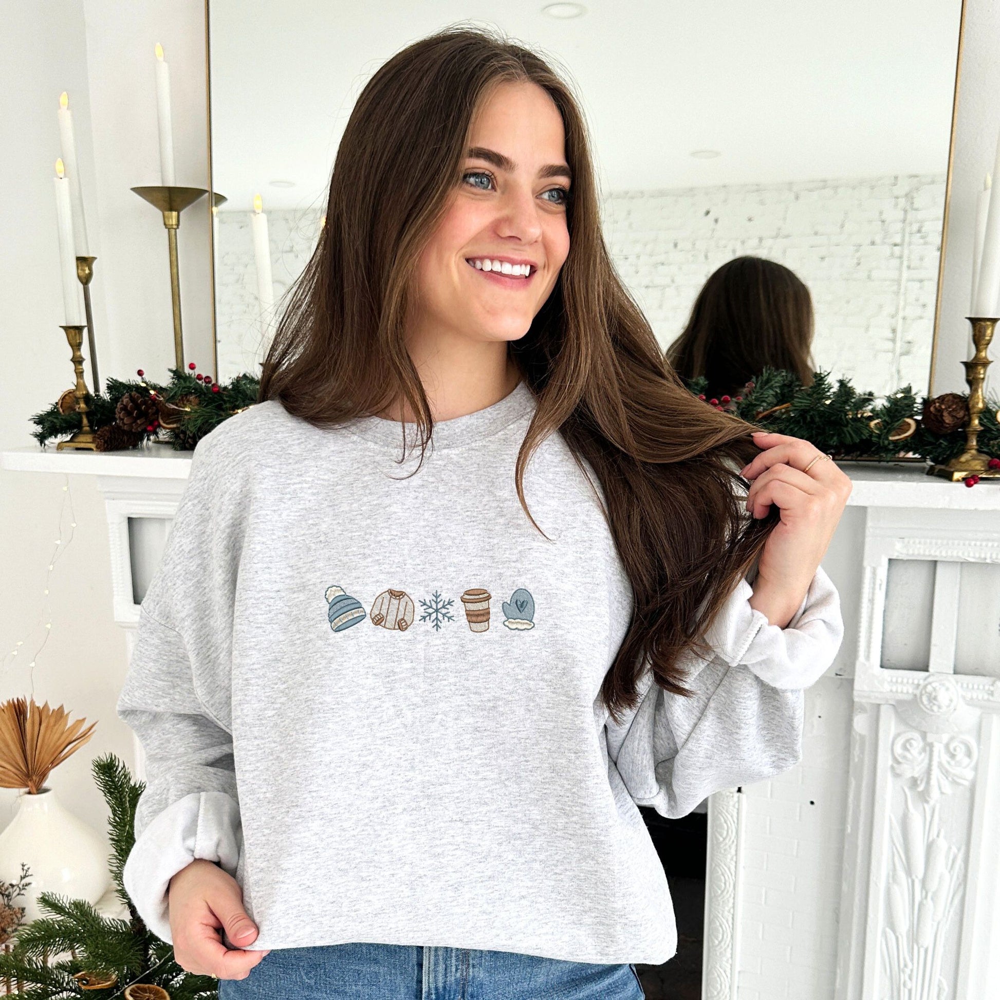 young woman wearing an ash crewneck sweatshirt with custom winter themed icons embroidered on the center chest. design featuring a cute beanie, sweater, snowflake, to-go coffee cup, and mitten in shades of blue, brown, and natural