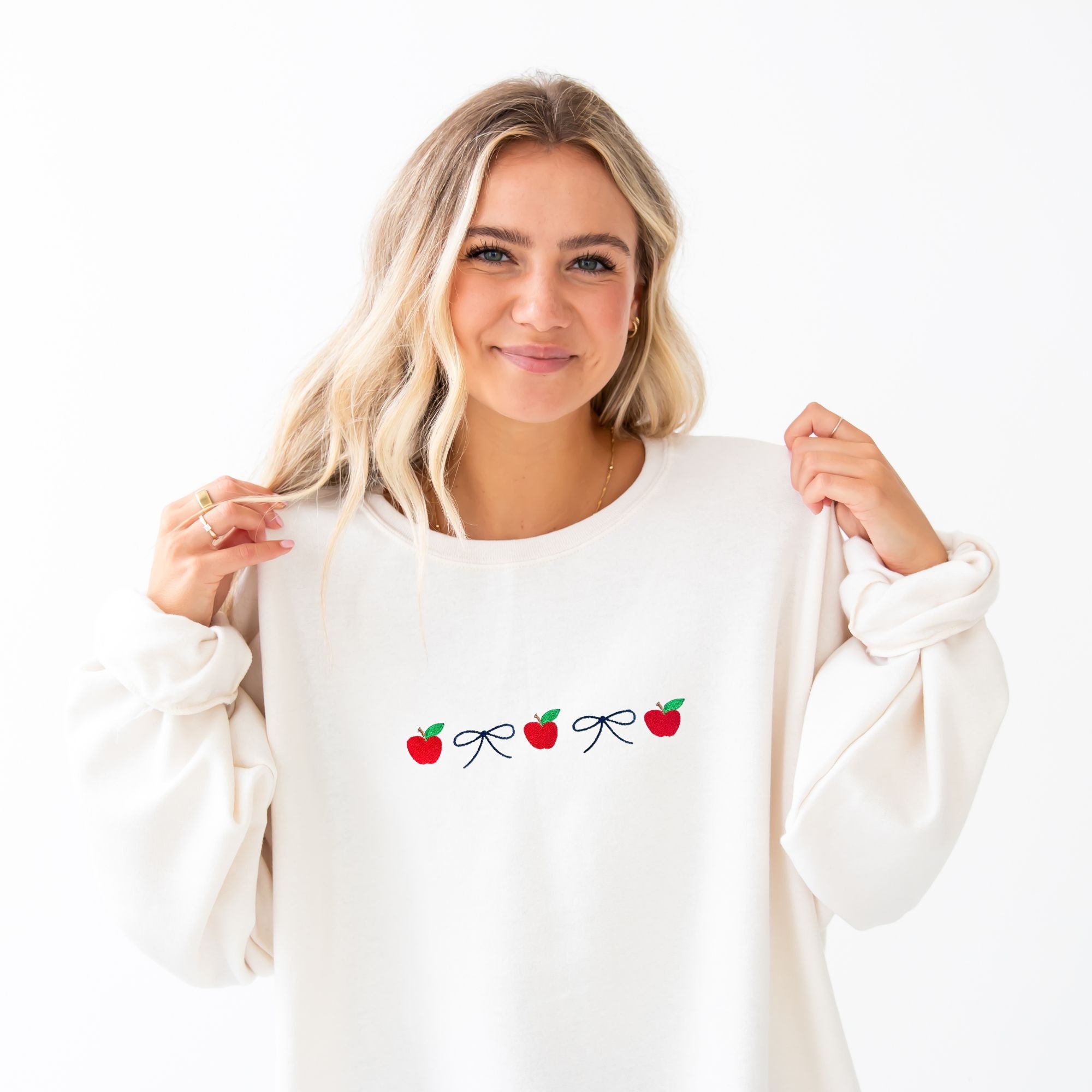 Woman wearing a white sweatshirt with red apple and bow embroidered design across the chest in an alternating pattern