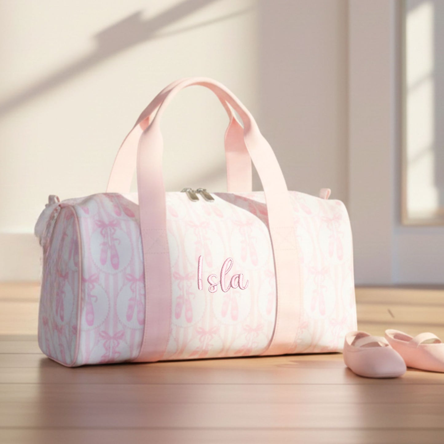 Pink duffel bag with ballet slipper design and 'Isla' monogram on a wooden floor.