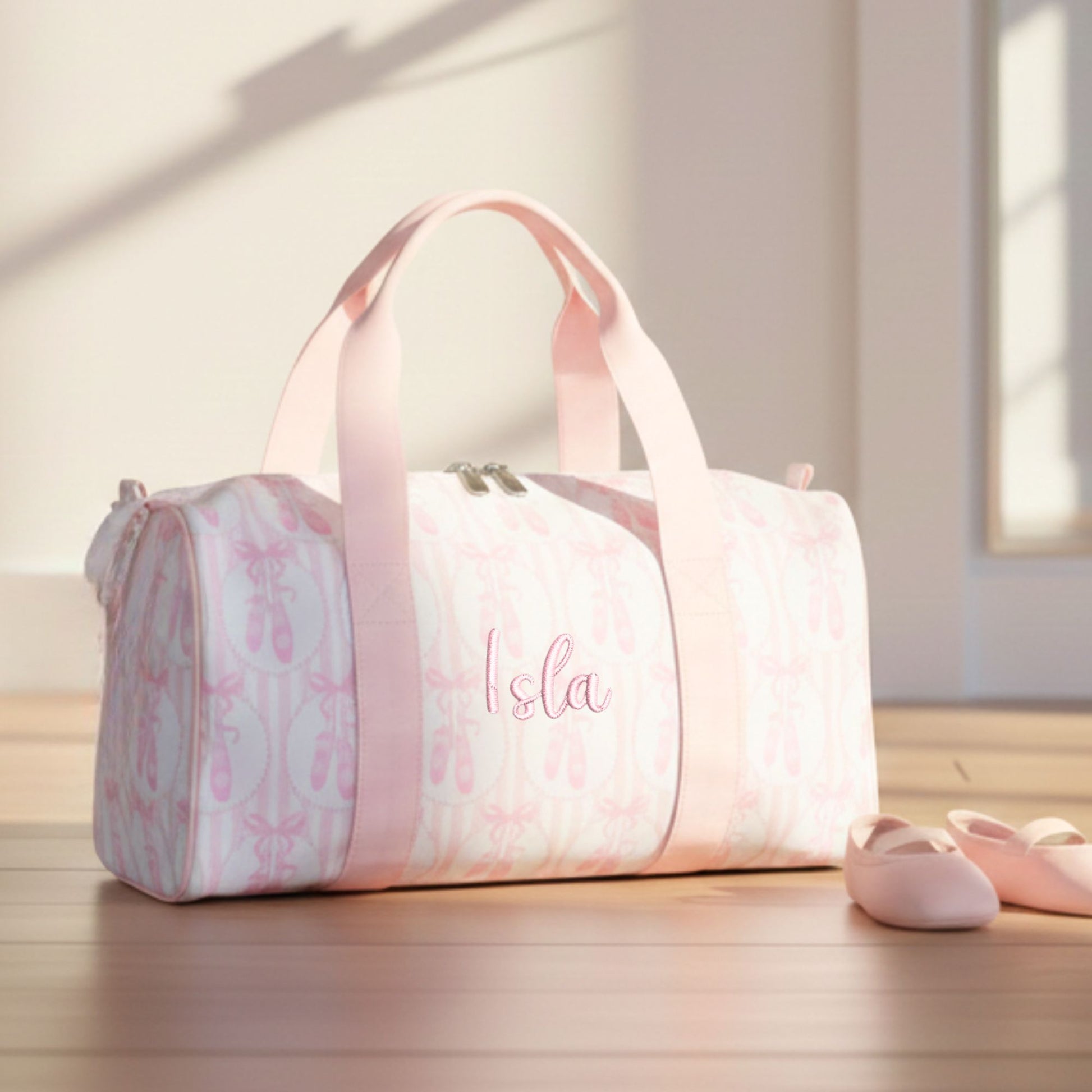Pink duffel bag with ballet slipper design and 'Isla' monogram on a wooden floor.