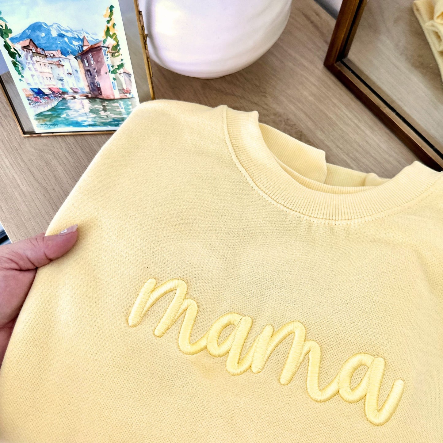 Yellow sweatshirt with 'mama' embroidery held by a hand on a wooden surface.