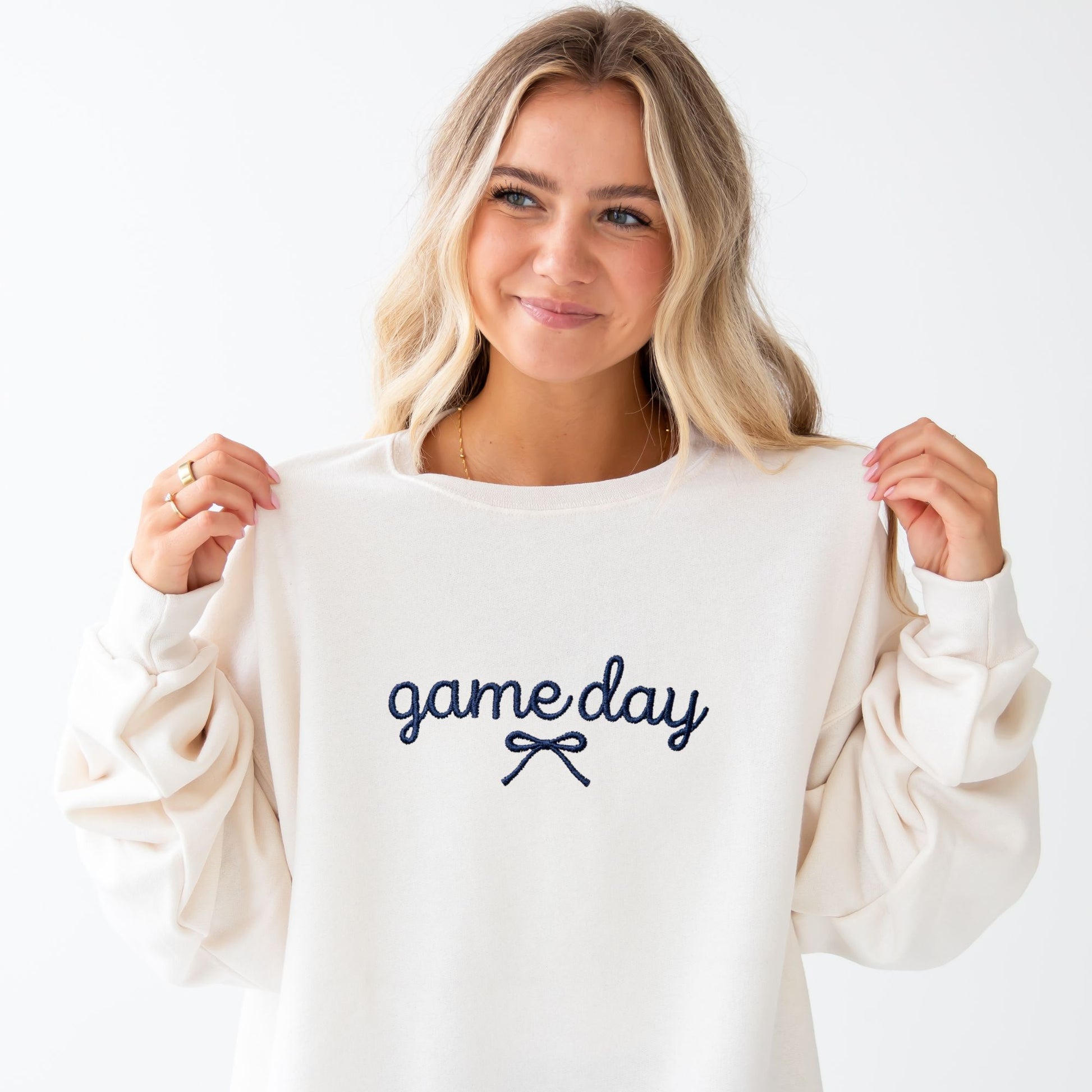A woman with blonde hair wearing a cream-colored crewneck sweatshirt with 'game day' embroidered in navy blue on the chest, holding the shoulders of the sweatshirt against a plain white background.