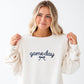 A woman with blonde hair wearing a cream-colored crewneck sweatshirt with 'game day' embroidered in navy blue on the chest, holding the shoulders of the sweatshirt against a plain white background.