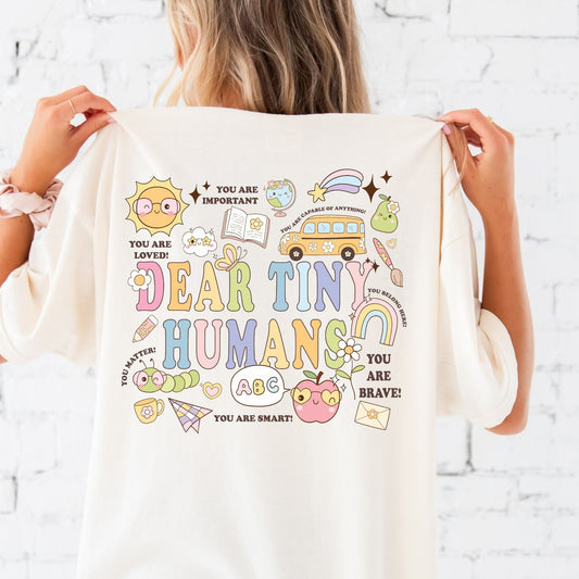 woman wearing an oversized comfort colors  t-shirt with colorful dear tiny humans print featuring colorful school/teacher icons and positive affirmations