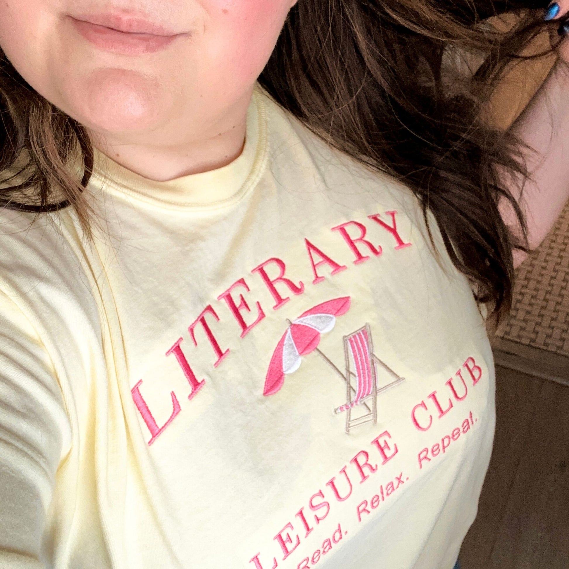 girl with brown hair wearing a banana comfort colors tee with a beach chair/umbrella and literary leisure club read relax repeat text embroidered design in pink and white thread. 