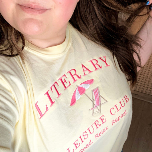 girl with brown hair wearing a banana comfort colors tee with a beach chair/umbrella and literary leisure club read relax repeat text embroidered design in pink and white thread. 