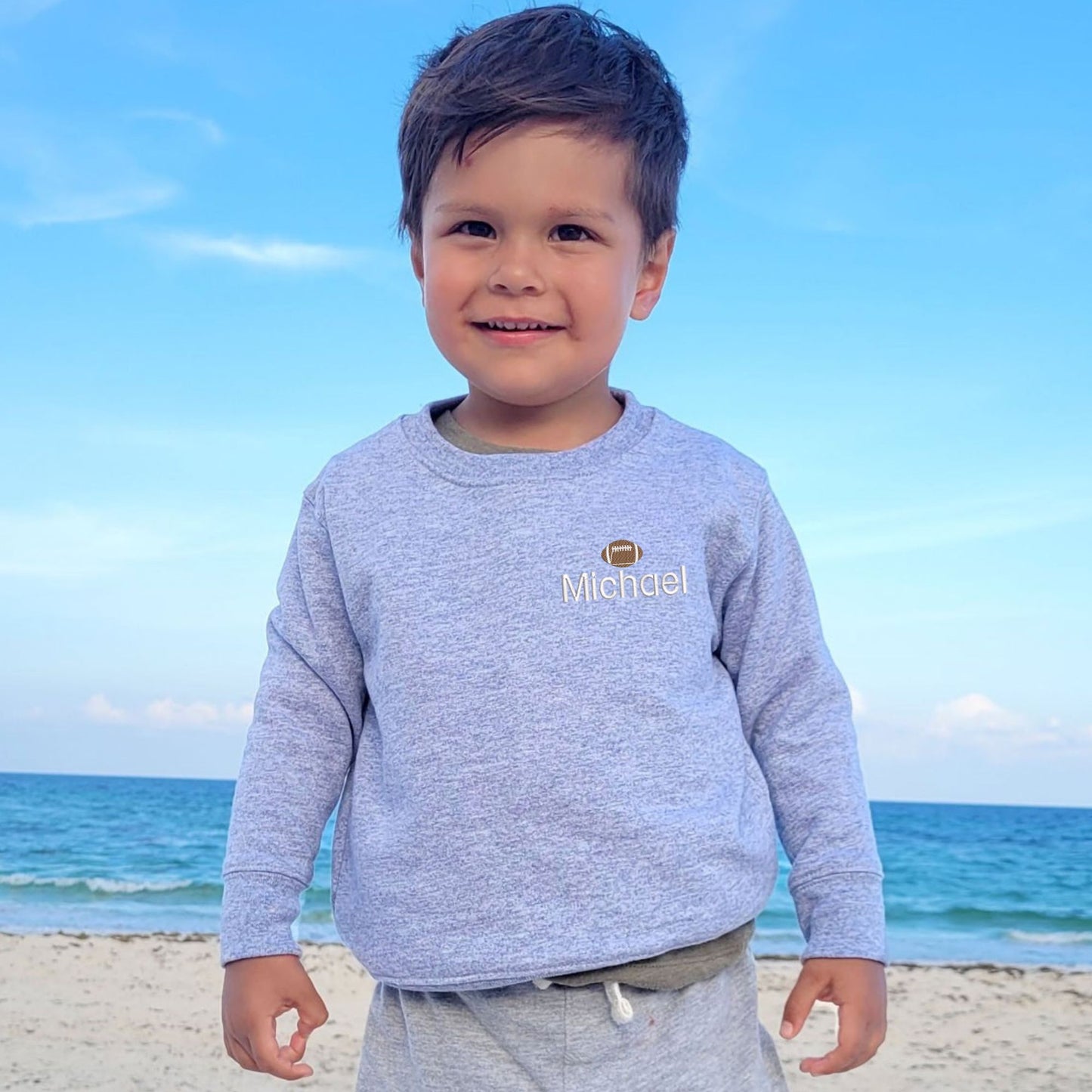 little boy wearing a heather gray sweatshirt with custom name embroidery on the left chest featuring a mini football above the name