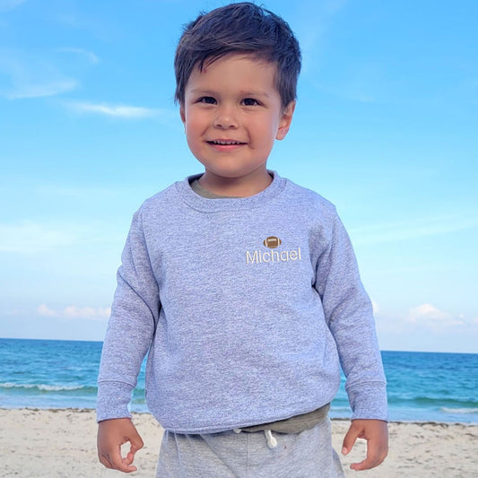 little boy wearing a heather gray sweatshirt with custom name embroidery on the left chest featuring a mini football above the name