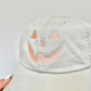 Ivory baseball cap with a pumpkin face embroidered design held up in front of a  light gray wall