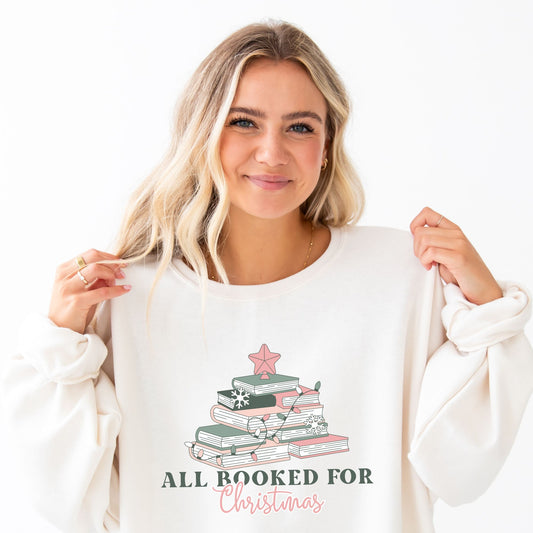 woman wearing an oversized sweet cream crewneck sweatshirt with a cute all booked for Christmas design featuring books stacked up to resemble a Christmas tree in shades of soft pink and greens