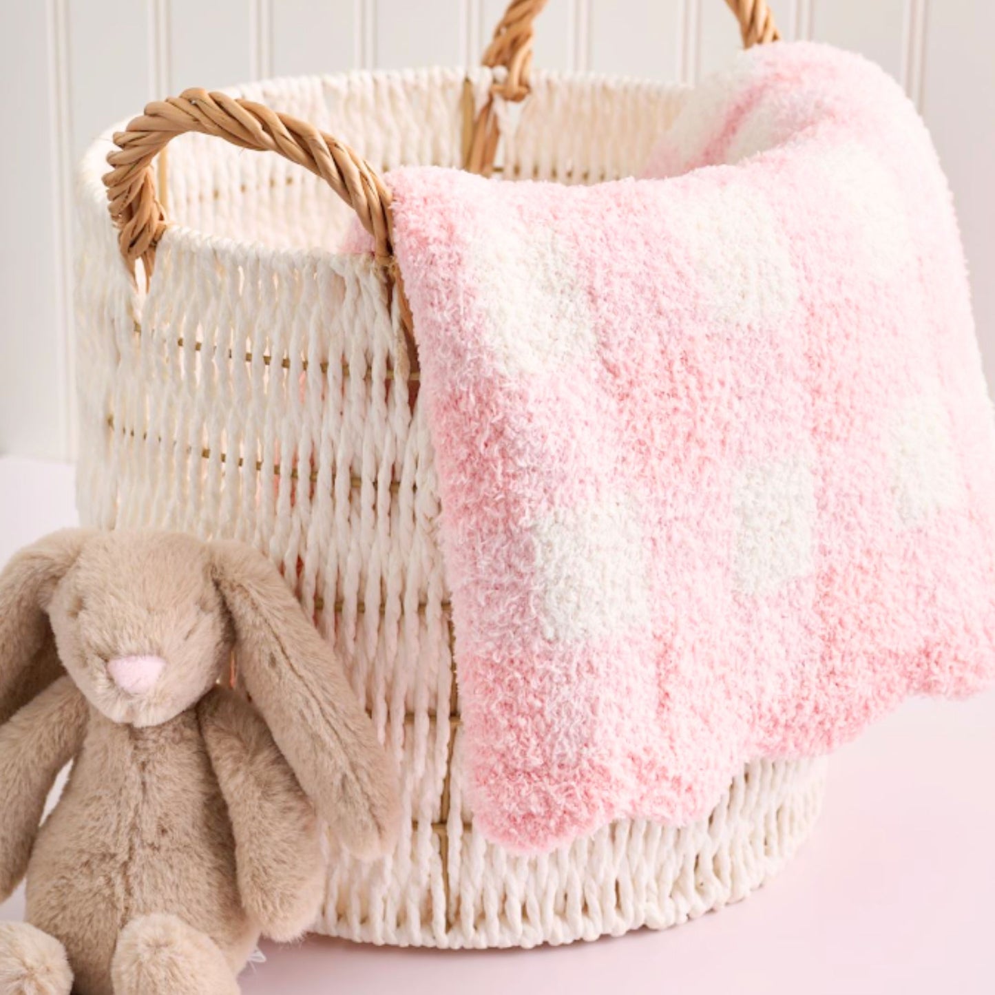 Wicker basket with pink and white blanket and brown plush bunny toy on a light background