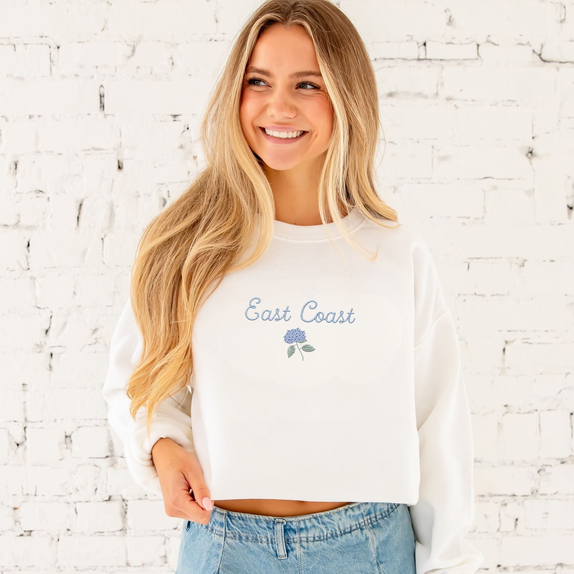 Woman wearing a white sweatshirt with 'East Coast' text against a white brick wall.