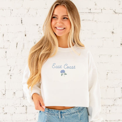 Woman wearing a white sweatshirt with 'East Coast' text against a white brick wall.