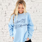 girl wearing a light blue crewneck sweatshirt with a custom just married design featuring a heart locket with a custom name in the center and wedding rings tied with a bow. design is printed large on the center chest in navy ink