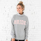 Woman wearing a gray sweatshirt with 'BRIDE' printed in pink on a white brick wall background