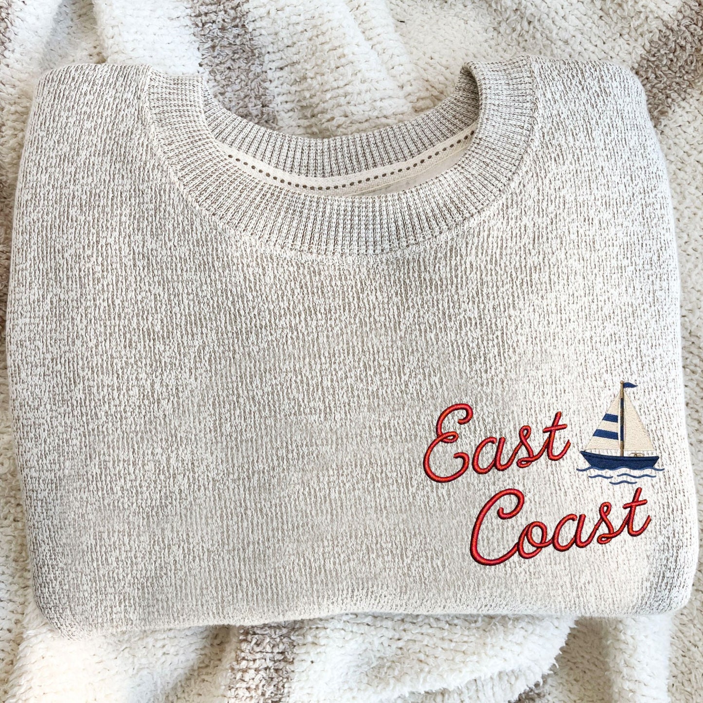 A folded light beige knit sweater with 'East Coast' embroidered in red script and a small blue-and-white sailboat motif, placed on a cream textured blanket.