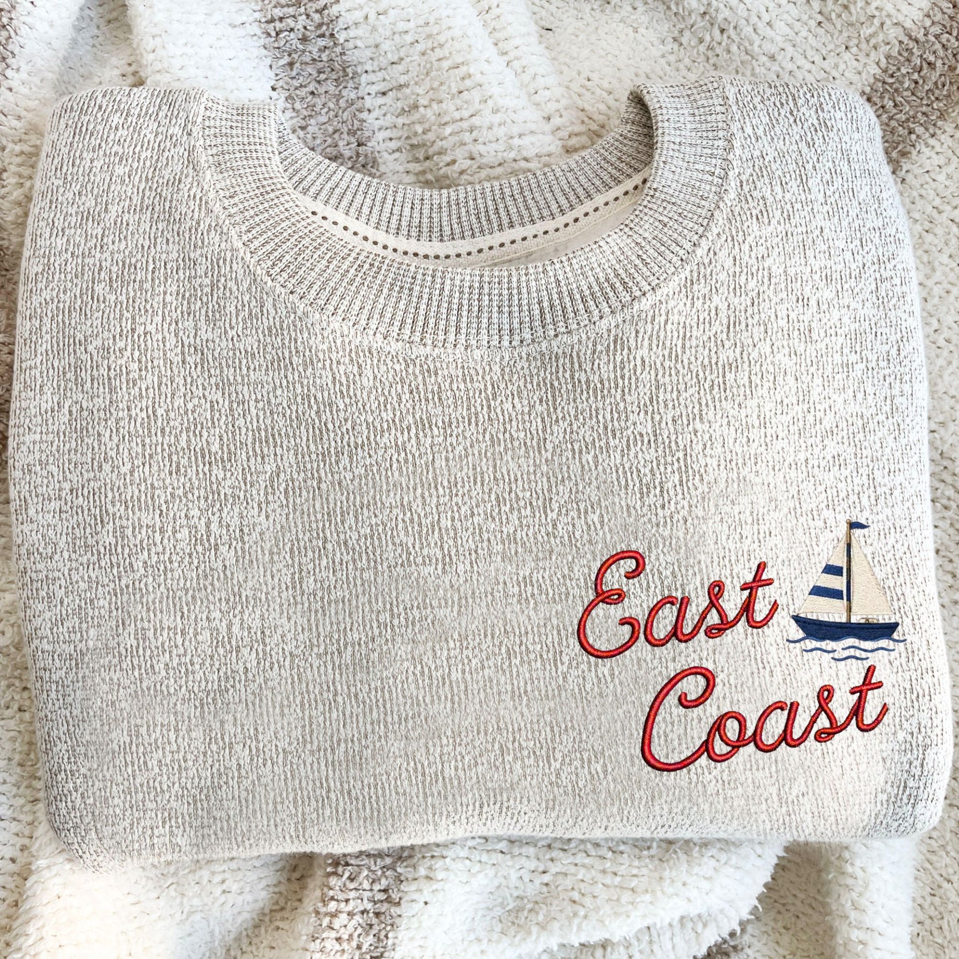 A folded light beige knit sweater with 'East Coast' embroidered in red script and a small blue-and-white sailboat motif, placed on a cream textured blanket.