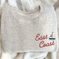 A folded light beige knit sweater with 'East Coast' embroidered in red script and a small blue-and-white sailboat motif, placed on a cream textured blanket.