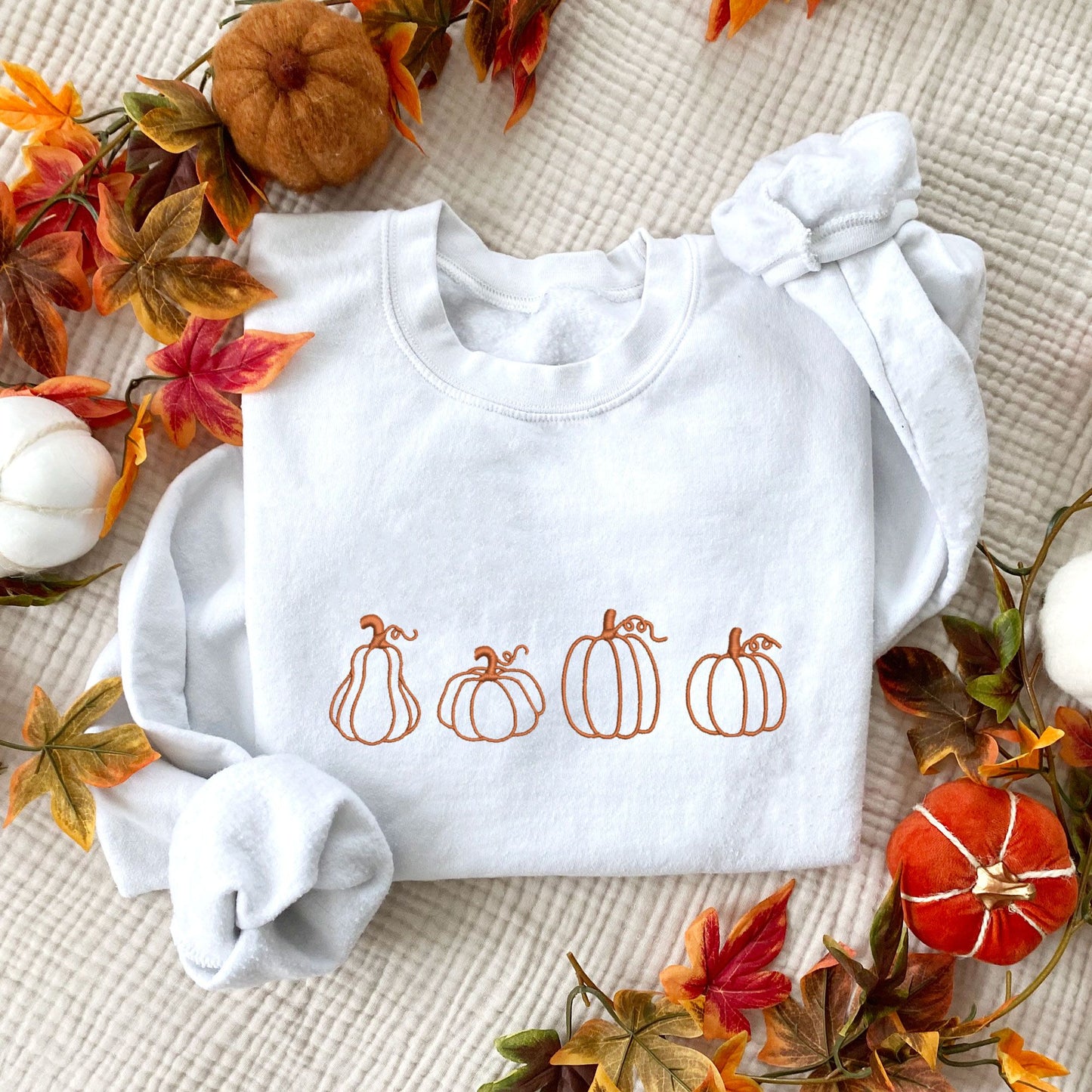 flatlay of a white crewneck sweatshirt with 4 different pumpkin outlines embroidered across the chest in pumpkin spice thread