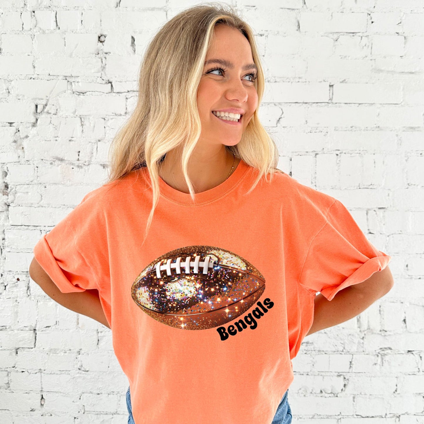 young woman wearing a melon comfort colors tee with large glitter football print and bengals text curved under the football in black ink