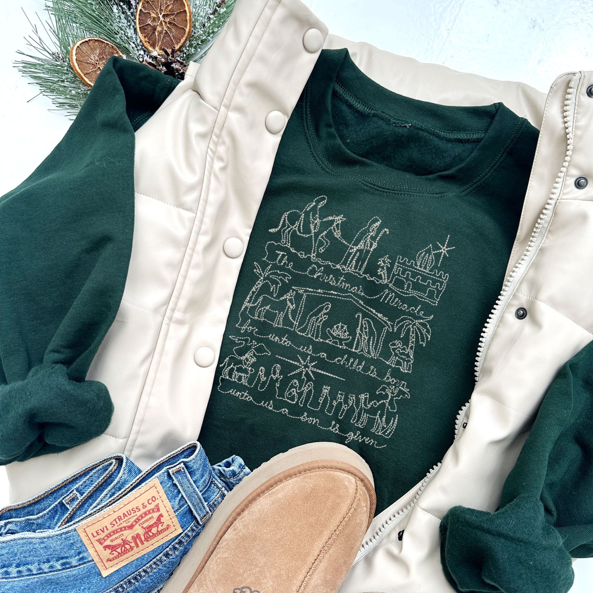 winter styled flat lay photo of a forest green crewneck sweatshirt with jeans and slippers and a vest. Embroidered on the chest of the sweatshirt is a stitched drawing of the Christmas nativity in natural thread.