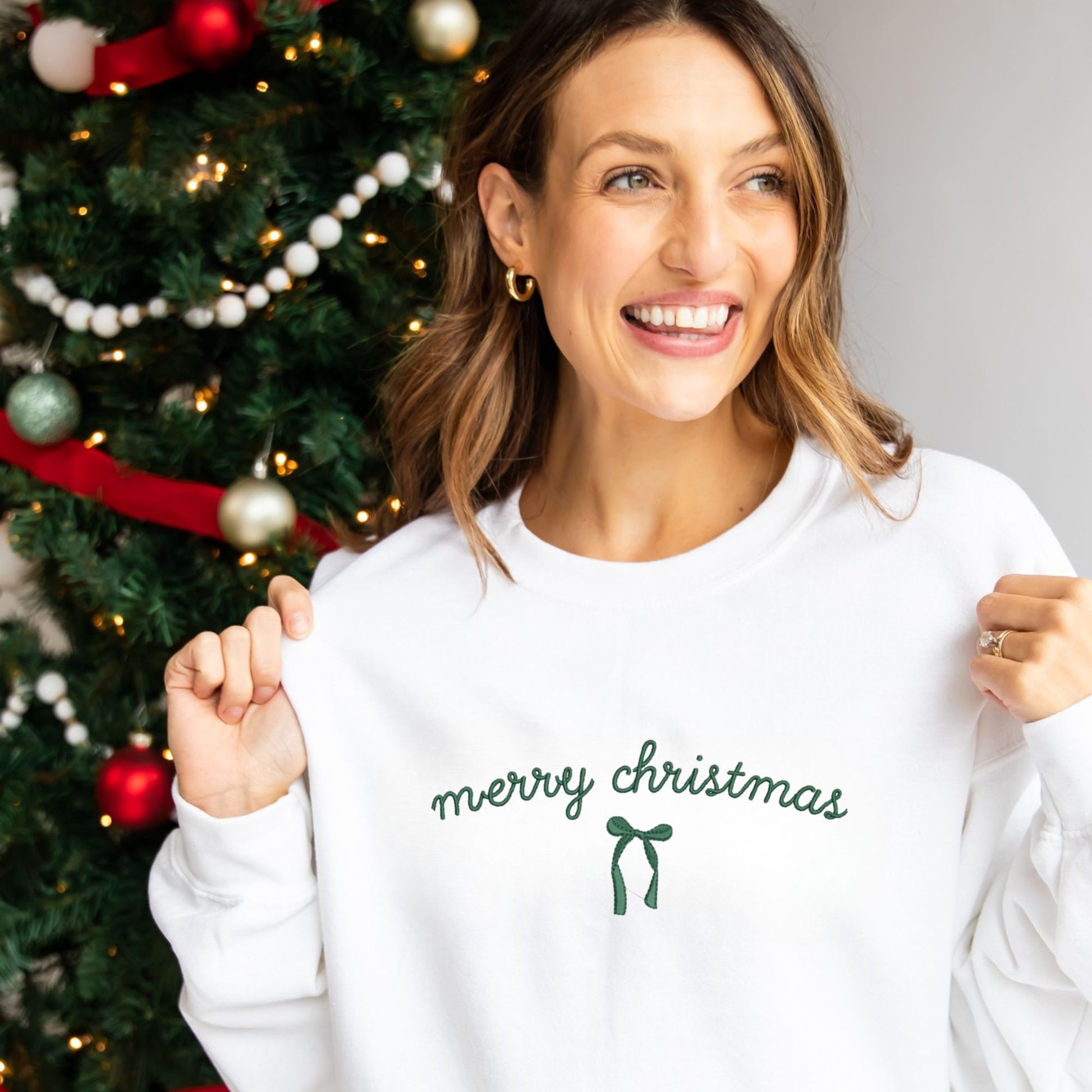 Woman wearing a 'merry christmas' sweater in front of a decorated Christmas tree.
