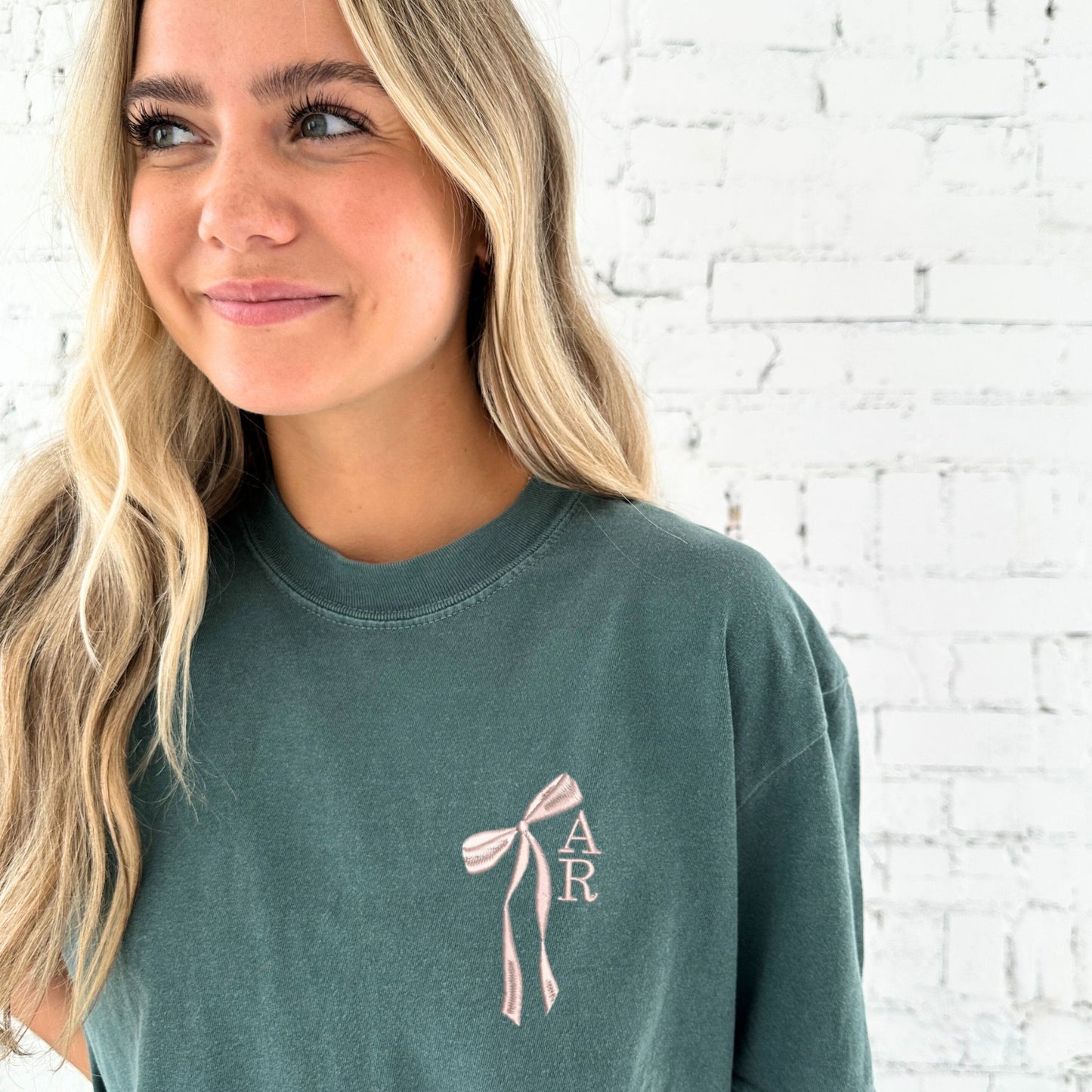 girl wearing a blue spruce comfort colors t-shirt with custom ribbon bow stacked initial embroidered design on the left chest in powder blue thread.