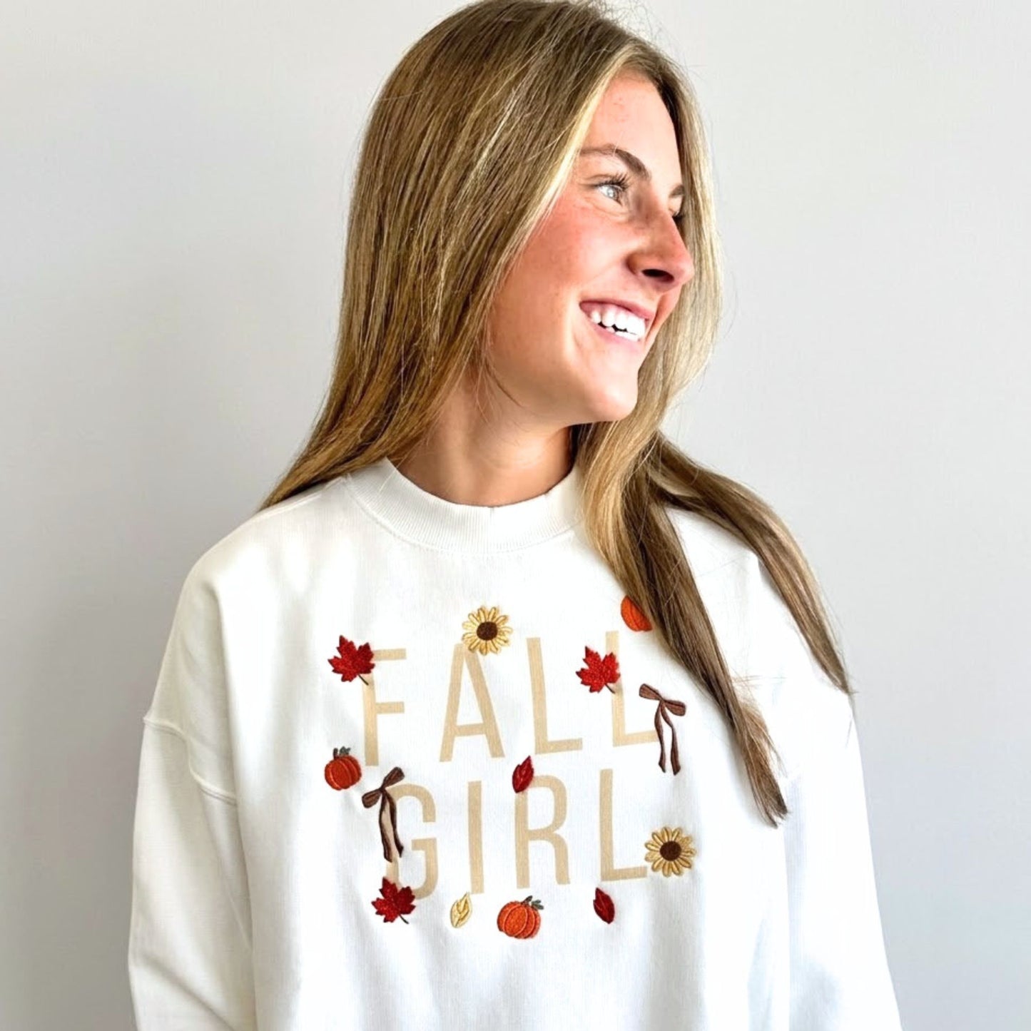 A woman with long blonde hair is shown from the chest up, wearing a white crewneck sweatshirt with a colorful 'FALL GIRL' graphic featuring autumn leaves, sunflowers, and pumpkins. She is smiling and looking to the side against a plain light gray background.