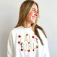 A woman with long blonde hair is shown from the chest up, wearing a white crewneck sweatshirt with a colorful 'FALL GIRL' graphic featuring autumn leaves, sunflowers, and pumpkins. She is smiling and looking to the side against a plain light gray background.