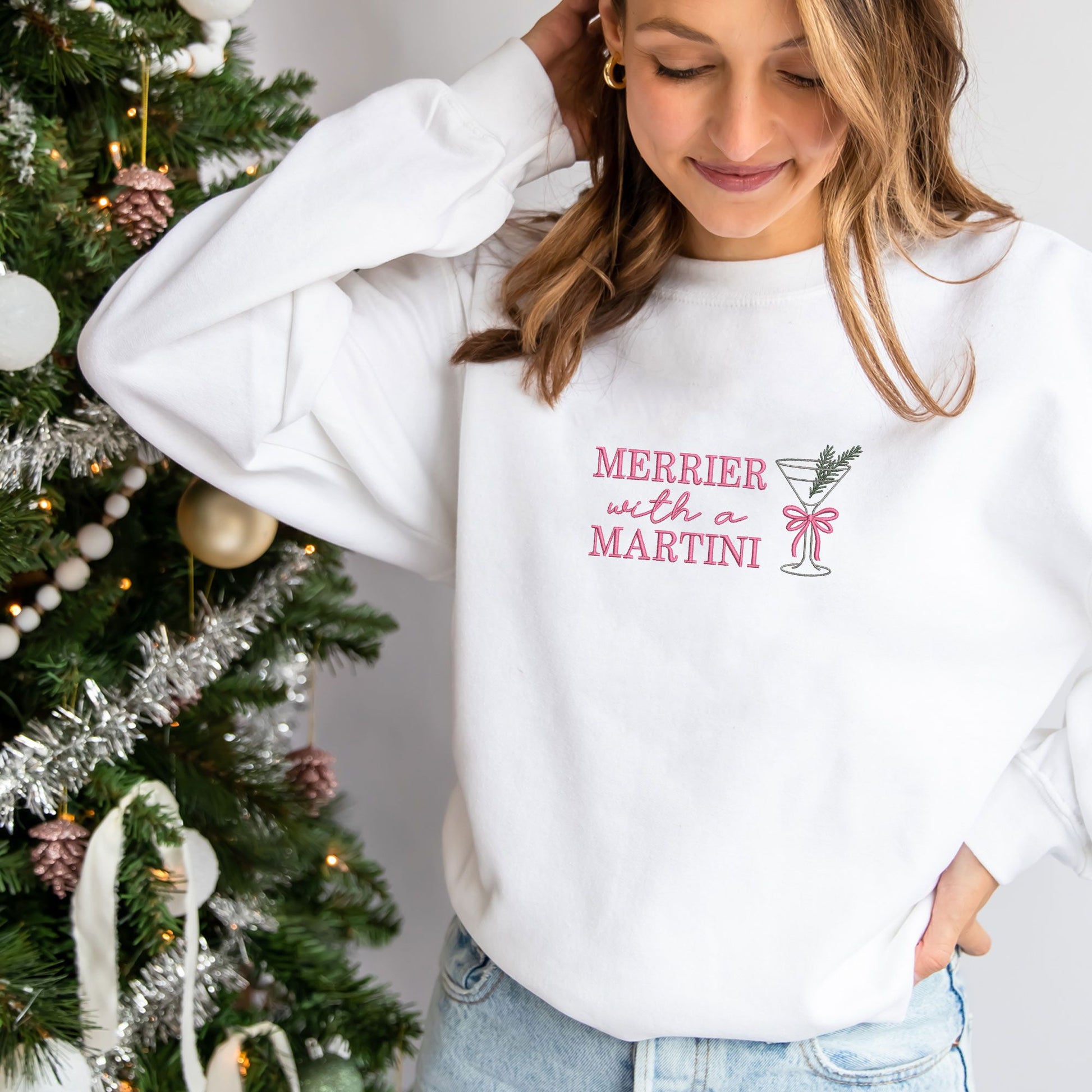 woman wearing a white crewneck sweatshirt with embroidered merrier with a martini text in pink thread and martini glass design with bow and pine sprig across the chest