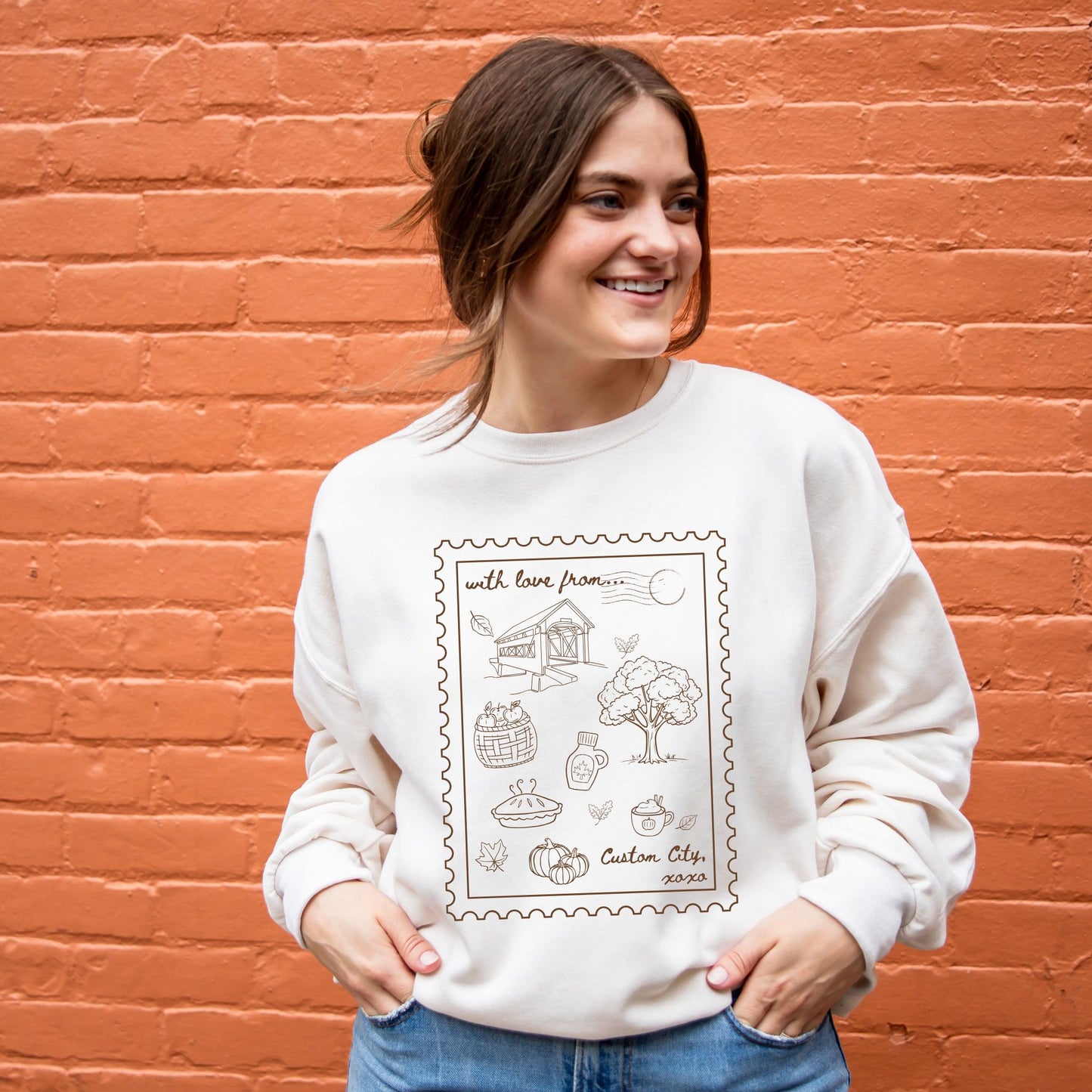 A woman wearing a cream-colored crewneck sweatshirt with a brown stamp-style graphic on the front, standing against an orange brick wall.