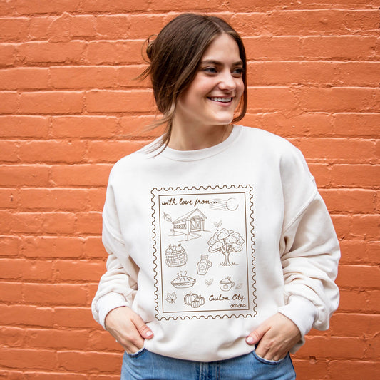 A woman wearing a cream-colored crewneck sweatshirt with a brown stamp-style graphic on the front, standing against an orange brick wall.