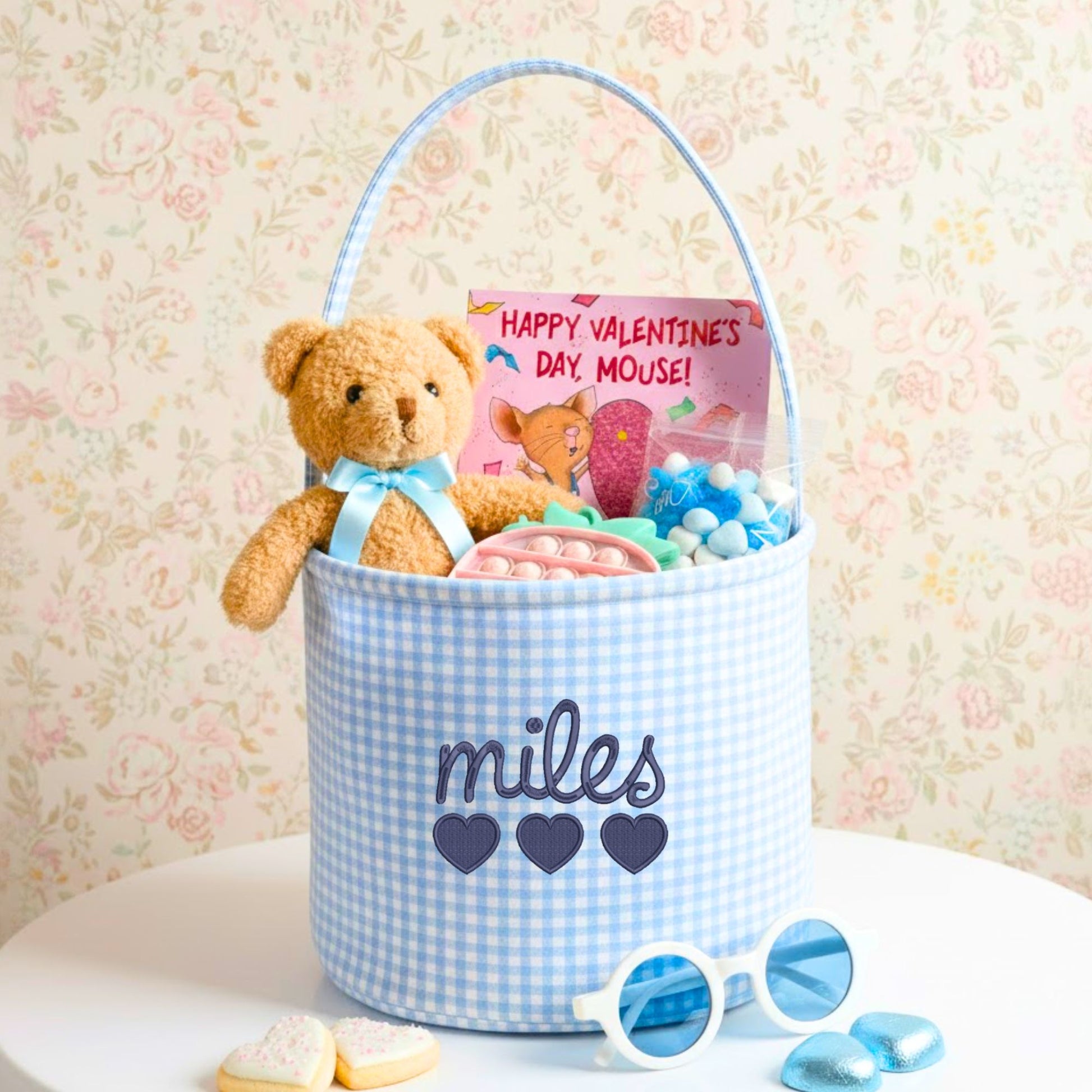 Blue gingham valentines gift basket with custom name and heart embroidery, stuffed with a teddy bear, candy, and 'Happy Valentine's Day Mouse!' book on a floral background.