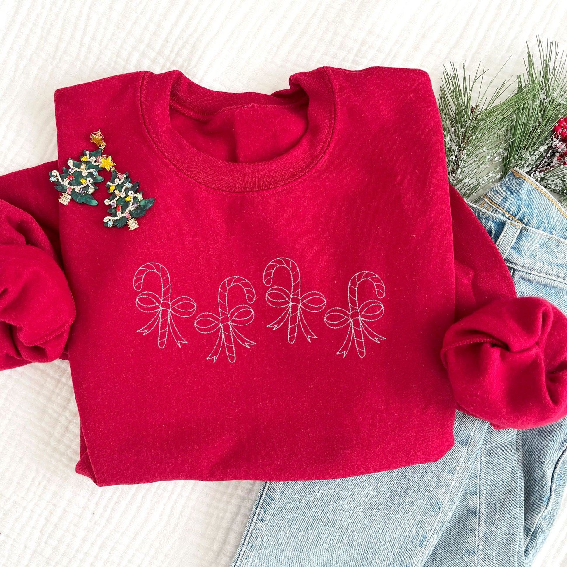 flat lay of a red crewneck sweatshirt with4 staggered outlined candy canes with bows across the chest in white thread.