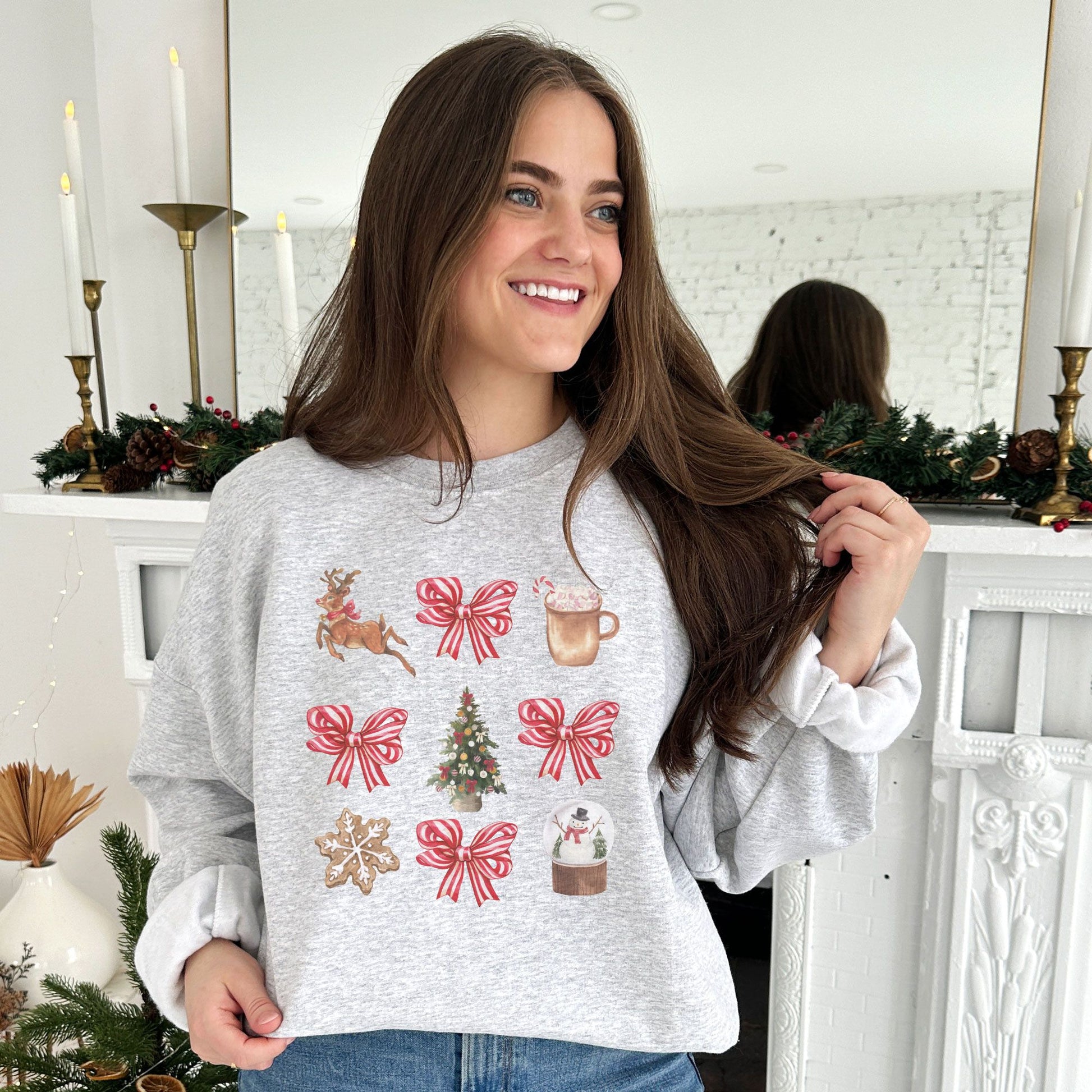 woman standing in front of a fireplace wearing an ash pullover with a retro reindeer, bow, cocoa, tree, cookie, and snow globe printed design