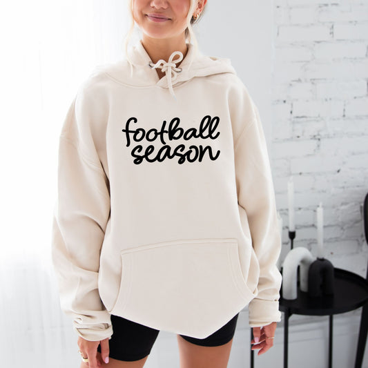 girl wearing a sandshell hooded sweatshirt with football season cursive black print