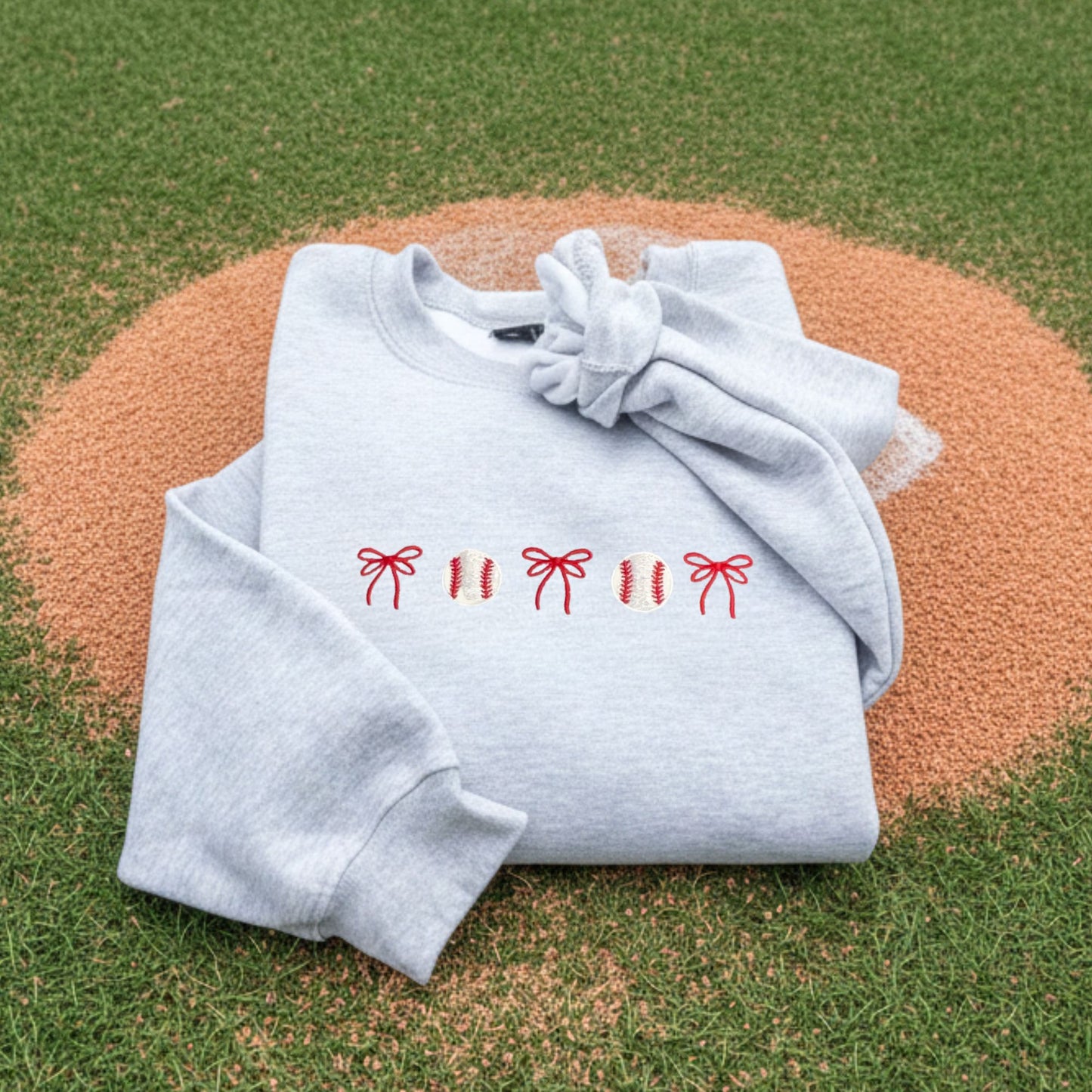 Folded light gray sweatshirt with embroidered baseball and bow design embroidered on the center chest on a baseball mound