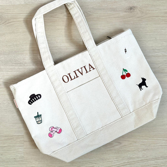 White tote bag with embroidered designs and 'OLIVIA' text on a wooden surface