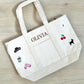 White tote bag with embroidered designs and 'OLIVIA' text on a wooden surface