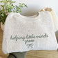 latte cozy knit nantucket fleece sweatshirt folded and held out above a light wooden dresser. The sweatshirt features a cute embroidered helping little minds grow design in a script font with a small delicate bow underneath. Embroidery is in eucalyptus thread.